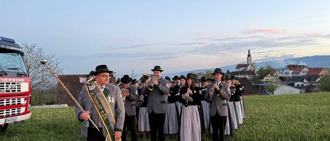 Eine Gruppe von Menschen in traditioneller Kleidung steht auf einer Wiese mit Musikinstrumenten. Ein Mann vorne hält einen Taktstock und eine Schärpe. Ein Feuerwehrauto ist auf der linken Seite geparkt.