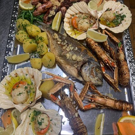 Ein silbernes Tablett zeigt eine Auswahl an Meeresfrüchten, darunter einen ganzen Fisch, Jakobsmuscheln, Garnelen und Kartoffeln, die mit Zitronenscheiben und Kräutern garniert sind.