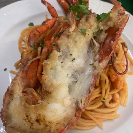 Ein Nahaufnahme eines Hummerschwanzes mit geschmolzenem Käse und Petersilie darauf, serviert auf einem Bett von Spaghetti mit roter Sauce.