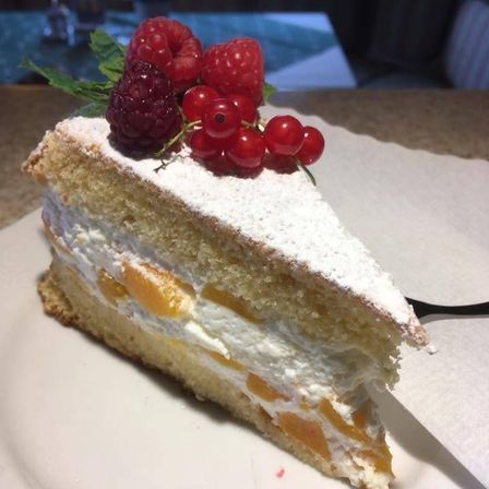 Ein Stück Kuchen mit cremiger Füllung, bestreut mit weißem Pulver, Himbeeren und roten Johannisbeeren auf einem weißen Teller.