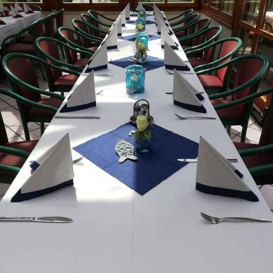 Ein langer Esstisch, der für eine Mahlzeit vorbereitet ist, mit blauen und weißen Tischläufern, gefalteten Servietten und ordentlich angeordnetem Besteck. Der Tisch ist von grünen Stühlen umgeben.