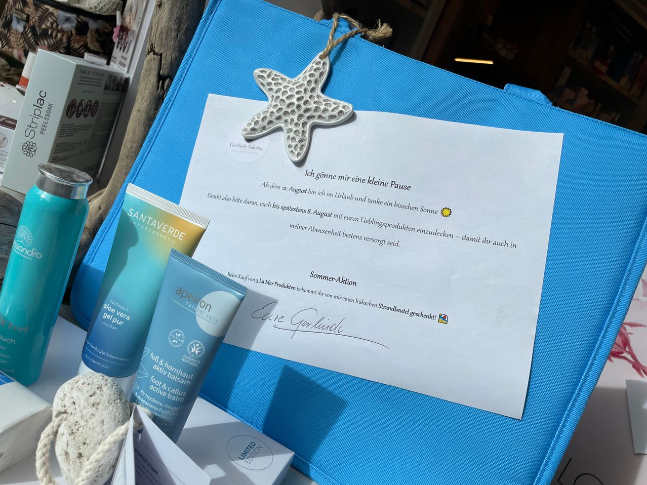 Ein handgeschriebener Zettel und einige Kosmetika in einer blauen Tasche. Der Zettel lautet: 'Ich gönne mir eine kleine Pause'. Darunter befindet sich eine handgeschriebene Unterschrift.