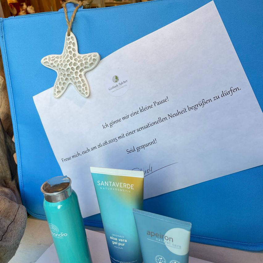 Ein weißes Papier mit einem Seestern-Ornament und deutschem Text liegt auf einer blauen Tasche. Darunter stehen eine grüne Flasche und zwei blaue Tuben auf einer weißen Oberfläche.