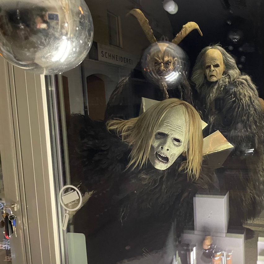 Eine Ausstellung von drei Halloween-Kostümen in einem Geschäft, darunter eines mit einer weißen Maske und einem Buch in der Hand, mit einem Schild darüber, das 'SCHNEIDEREI' steht.