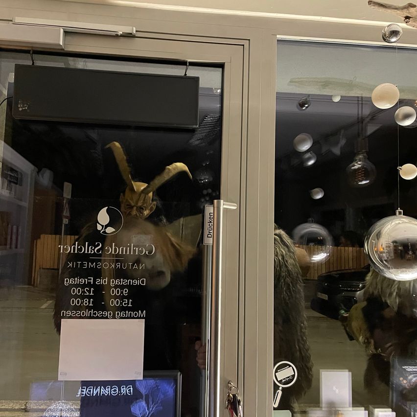 Ein Ladentür mit einem Schild, das 'Geschlossen heute' anzeigt. Drinnen ein goldener Horn und ein Zettel. Die Scheibe reflektiert ein gehörntes Wesen und Lichter.