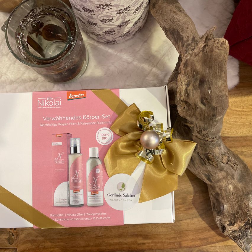 Eine pinke und weiße Geschenkbox mit einer goldenen Schleife steht auf einem Holztisch. Die Box ist von Demeter und trägt die Marke Nikolai. Sie enthält ein Set an Körperpflegeprodukten.
