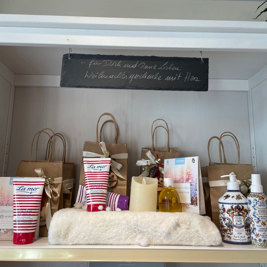 Ein weißer Schrank zeigt ein Tafel mit deutschem Text, mehrere Geschenktüten, eine Kerze, Körperlotion und Handseifeflaschen. Eine weiße Pelzdecke ist im unteren Regal.
