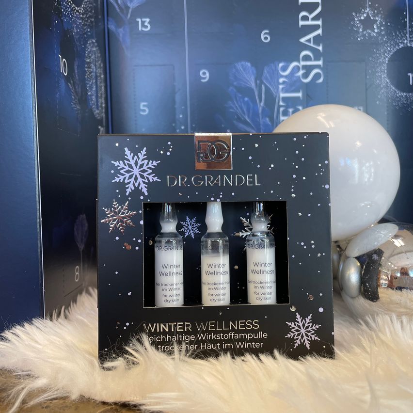 Eine Schachtel mit drei kleinen Flaschen der Dr. Grandel Winter Wellness Hautpflegeprodukte, auf einer weißen Pelzoberfläche mit Schneeflocken darauf platziert.