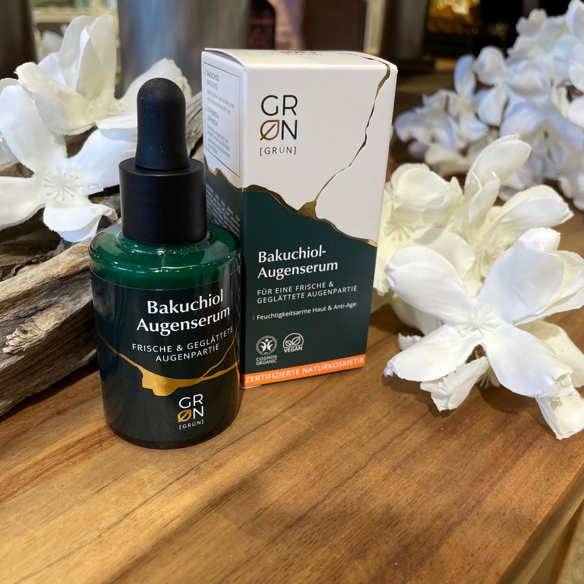 A green bottle of Bakuchiol Eye Serum is placed on a wooden table with its box. The box is labeled with 'GR ON' and is green and white. White flowers surround the table.