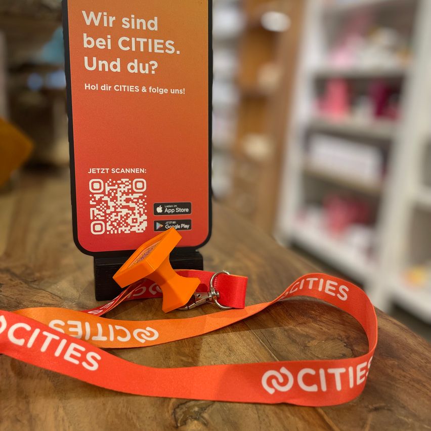 Ein orangefarbener Lanyard mit dem Text 'CITIES' liegt auf einem Holztisch. Dahinter befindet sich ein orangefarbenes Schild mit dem Text 'Wir sind bei CITIES. Und du?' und einem QR-Code.