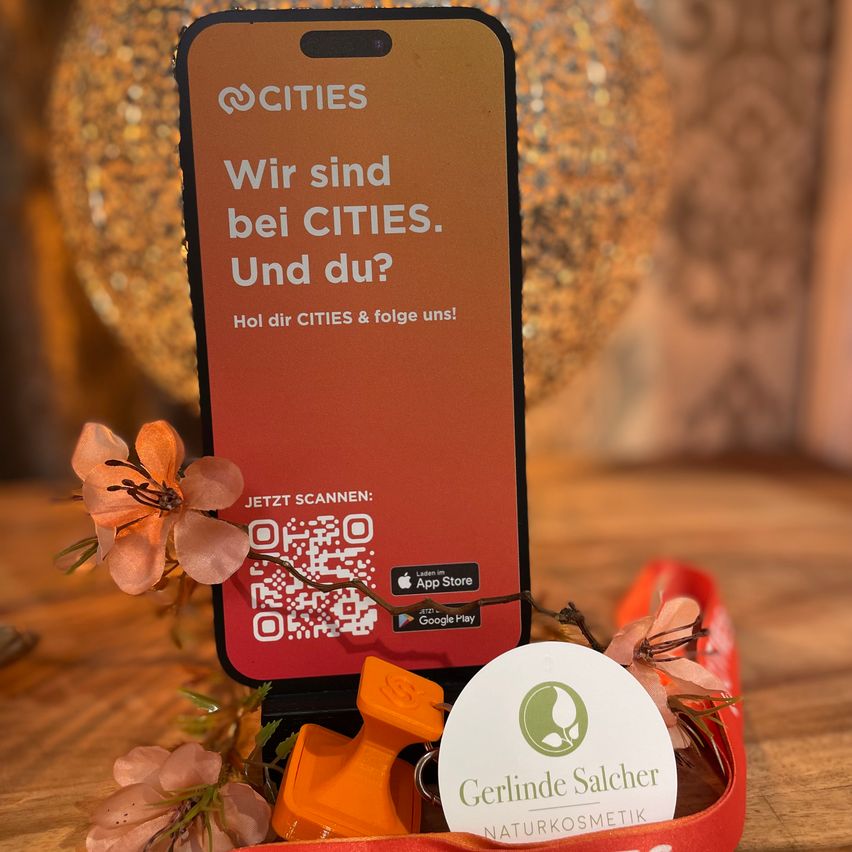 Ein Smartphone zeigt einen roten Hintergrund mit weißem Text, der 'Wir sind bei CITIES. Und du? Folge uns!' lautet, darunter ein QR-Code. Es steht auf einem Tisch mit Blumen und einem Anhänger.