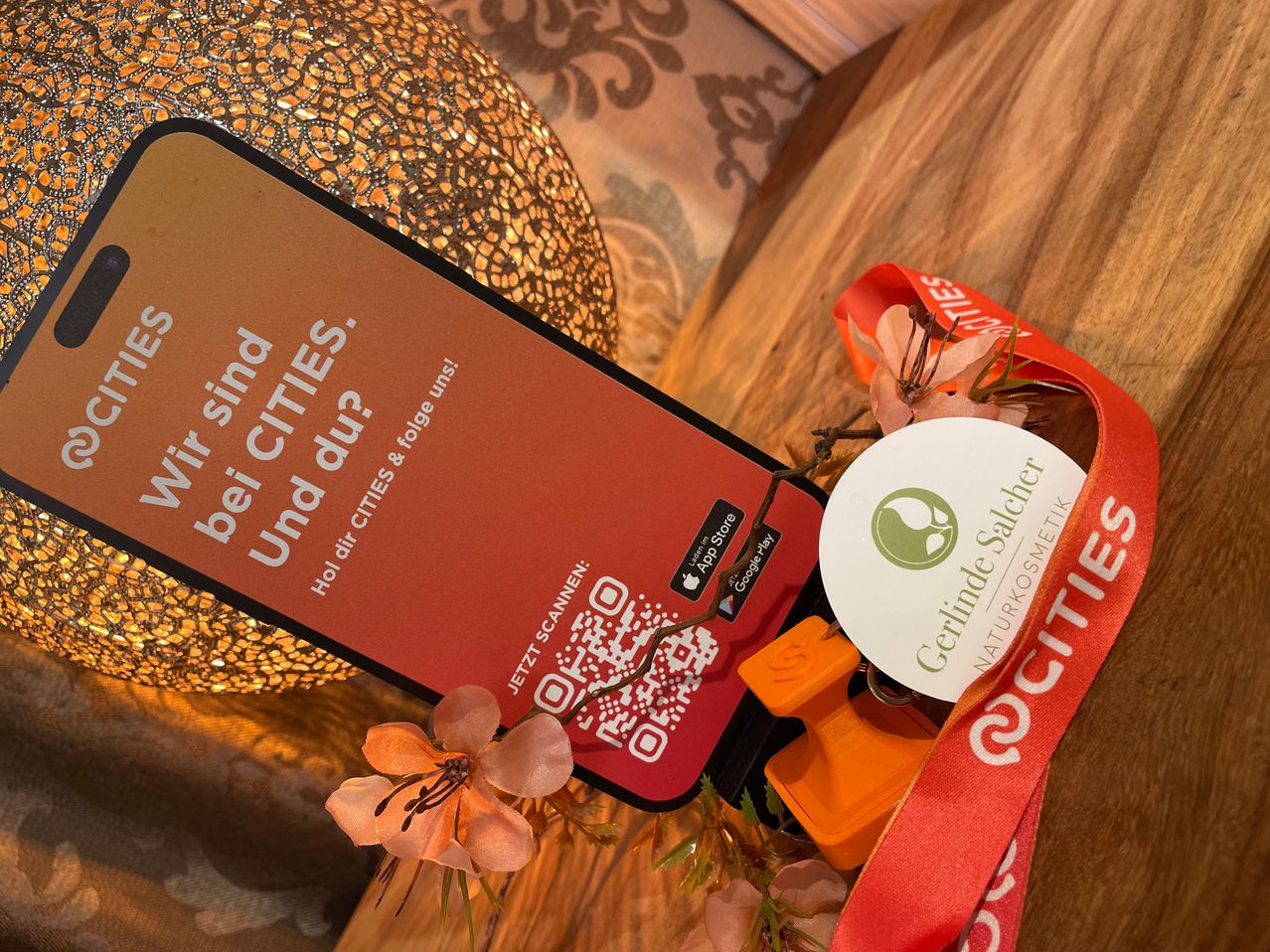 Ein Smartphone mit rotem Hintergrund zeigt den Text 'Bist du mit Städten?'. In der Nähe befindet sich ein rotes Band, ein weißer runder Anhänger mit Logo und einige Blumen.