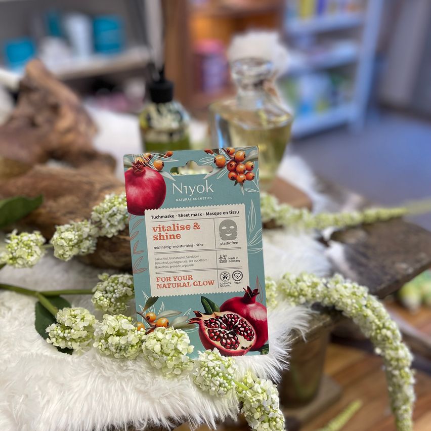 Eine Schachtel von Niyoks natürlichem Glow-Gesichtsmasken-Set liegt auf einer pelzartigen Oberfläche. Die Schachtel zeigt eine Granatapfel und Blumen, die ihre natürlichen Inhaltsstoffe hervorheben.