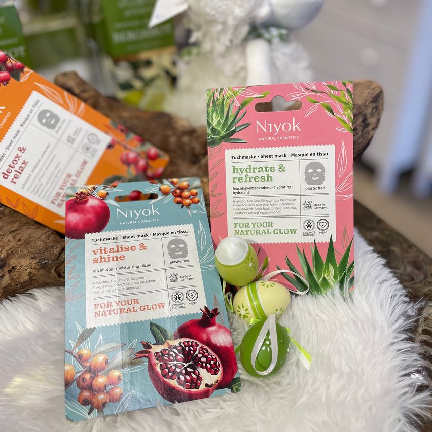 Zwei Niyok-Tuchmasken auf einer weißen Pelzoberfläche. Eine ist rosa mit Aloe Vera und die andere blau mit Granatapfelsamen.