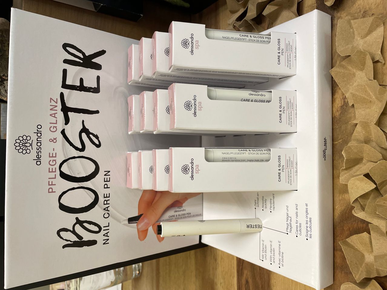 A display of four white and pink boxes of a nail care pen product labeled 'Booster' with a hand holding one of the pens.