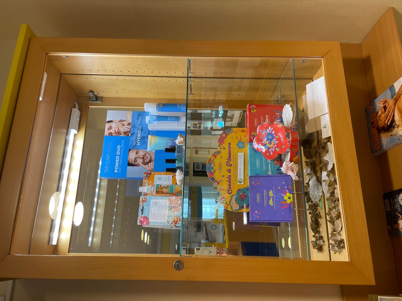 Eine Holzvitrine mit Glasfront zeigt verschiedene Artikel, darunter ein blaues Kästchen, bunte Schachteln und eine Karte mit einem Bild eines Mannes. Die Produkte sind ordentlich angeordnet, mit einem herzförmigen Kasten und einer weißen Box in der Ecke. Es gibt eine Reflexion einer Person und eines Lichts auf dem Glas.
