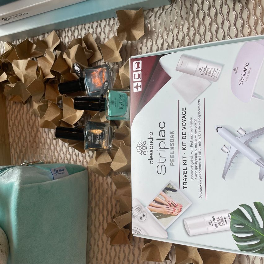 Eine Box des Striplac-Reisesets mit Fuß- und Handpflegeprodukten liegt auf einem Tisch mit Nagellackflaschen. Die Box hat eine Flugzeugillustration.