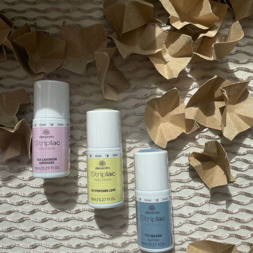 Drei vegane Striplac-Nagellackflaschen auf einem strukturierten Hintergrund. Eine ist Lavendel-Limonade, eine ist Popcorn-Liebe und eine ist Meeresglitzer.