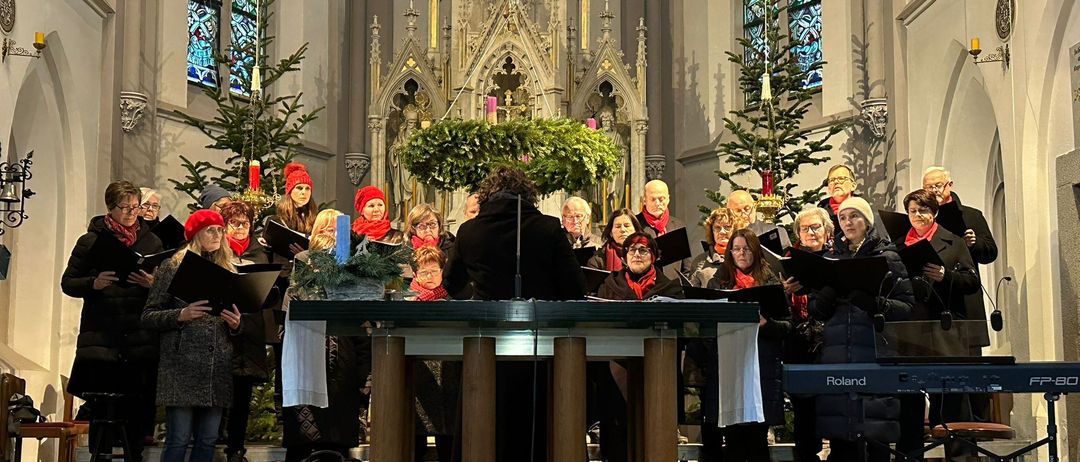 Ein Chor singt in einer Kirche, geschmückt mit Weihnachtsdekorationen, darunter Weihnachtsbäume und Kerzen. Die Chormitglieder halten ihre Notenblätter und stehen vor einem Klavier und einem Podium.