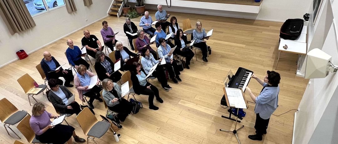Eine Gruppe von Menschen versammelt sich in einem Saal, sitzt auf Stühlen und hält Bücher. Eine Person steht vor einem Klavier und spielt es, während eine andere Person neben ihr steht.