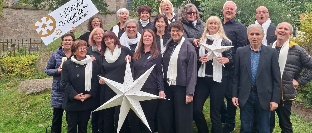 Eine Gruppe von Menschen steht im Freien in einem Park mit Bäumen und einer Steinmauer, lächelt und posiert für ein Foto. Sie tragen Winterkleidung und einige halten einen weißen Stern. Ein Schild steht 'Die Vielfalt des Advents erleuchten'.