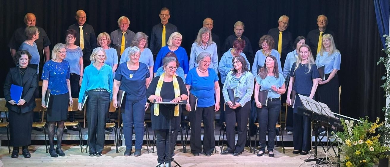 A choir of people stands on a stage, holding music sheets, with a black curtain in the background. They wear matching blue shirts and yellow ties.