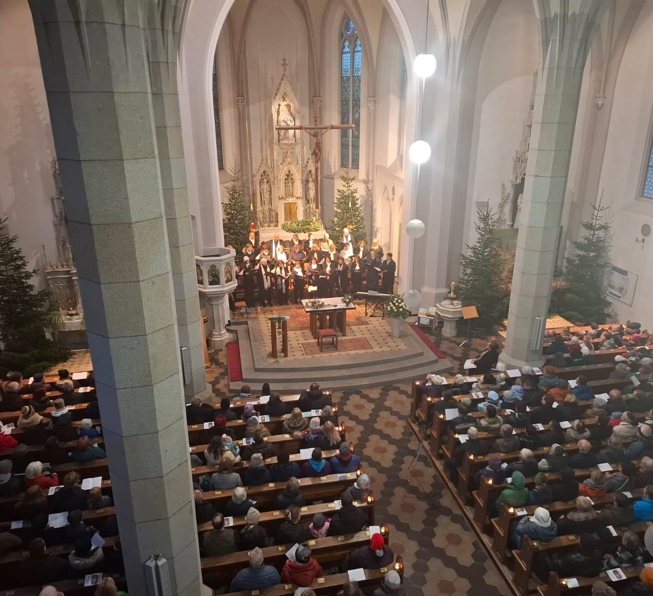 A choir performs in a church with an altar and Christmas trees. The audience sits on pews, holding candles. The church features stained glass windows and a high ceiling.