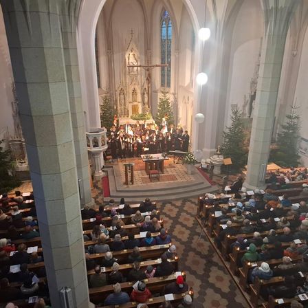 Ein Chor singt in einer Kirche mit Altar und Weihnachtsbäumen. Das Publikum sitzt auf Kirchenbänken und hält Kerzen. Die Kirche hat Buntglasfenster und eine hohe Decke.