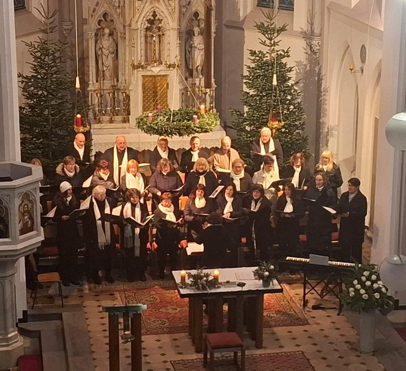 A choir in formal attire, singing in a church with Christmas decorations, candles, and musical instruments on a table. Statues and a pulpit are in the background.