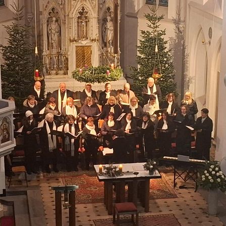 Ein Chor in formeller Kleidung singt in einer Kirche mit Weihnachtsdekoration, Kerzen und Musikinstrumenten auf einem Tisch. Im Hintergrund befinden sich Statuen und eine Kanzel.