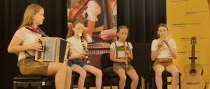 Vier junge Mädchen sitzen auf einer Bühne und spielen Musikinstrumente. Eine spielt Akkordeon, eine andere Flöte, und die anderen Klarinette und Harfe. Sie treten bei einer Veranstaltung auf.