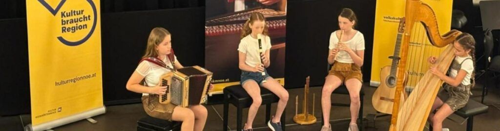 Drei Mädchen sitzen und spielen Instrumente. Eine spielt Akkordeon, eine andere Blockflöte und die dritte Harfe. Sie befinden sich auf einer Bühne mit einem Hintergrund, der Musikinstrumente zeigt.