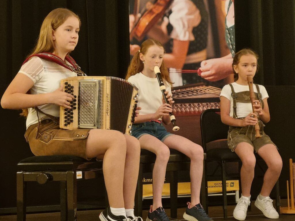 Drei Mädchen spielen Musikinstrumente bei einer Aufführung. Das Mädchen links spielt Akkordeon, die mittlere Flöte und die rechte Blockflöte. Sie sitzen auf Stühlen vor einem Poster.