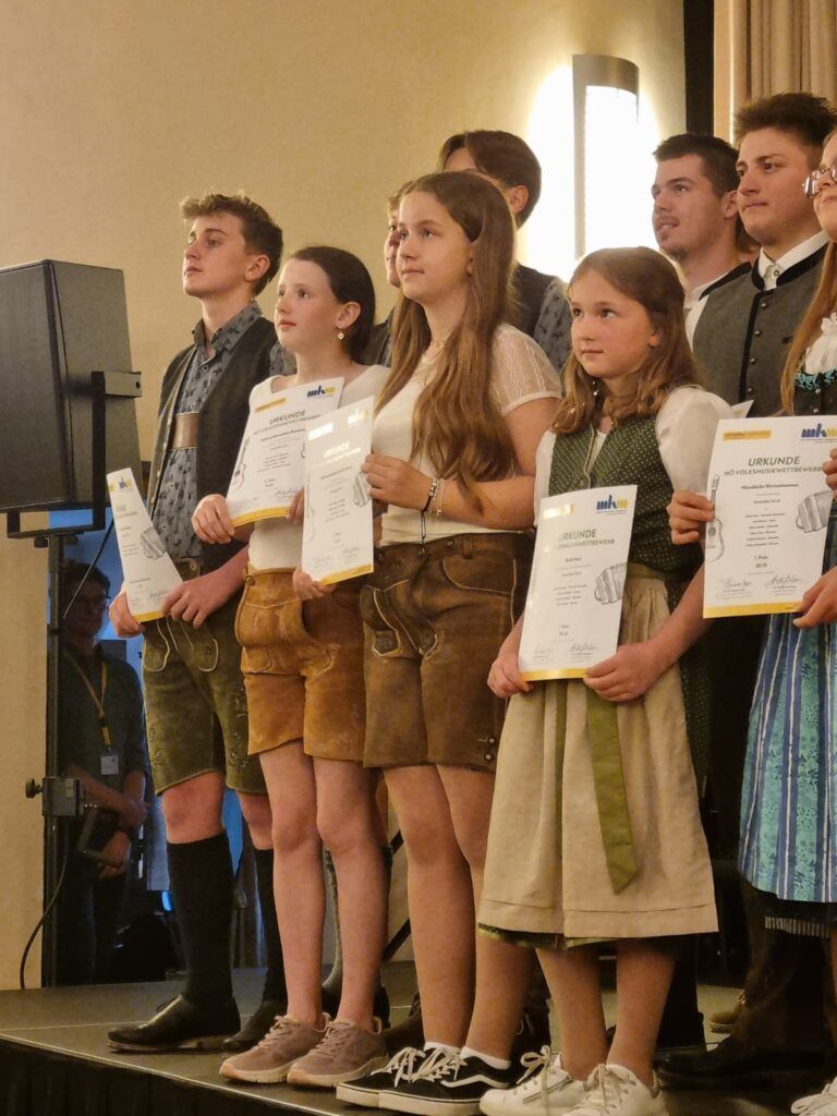 Eine Gruppe junger Menschen in traditioneller deutscher Kleidung, die Urkunden halten, steht zusammen in einem Raum. Ein Lautsprecher und eine Person mit Ausweis sind im Hintergrund zu sehen.