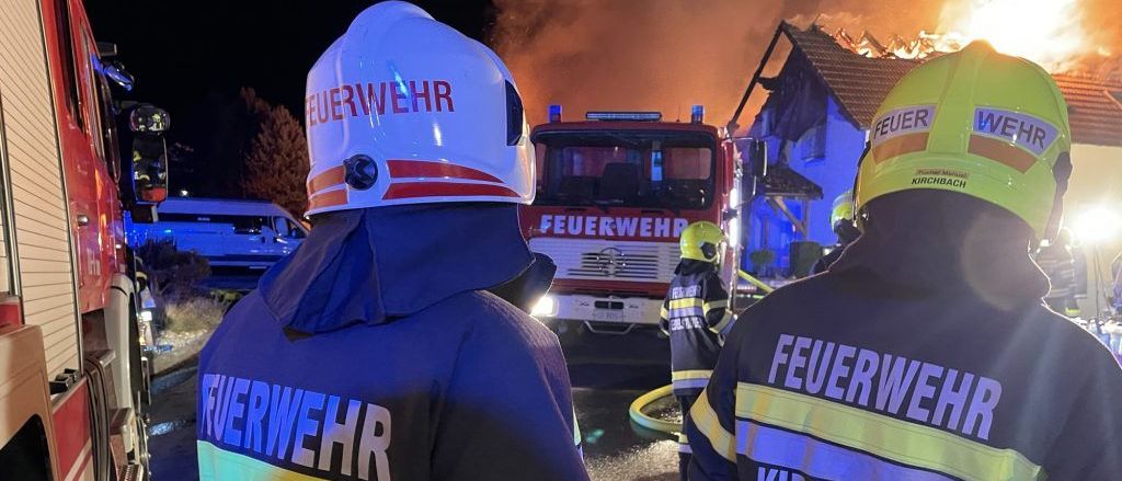 Feuerwehrleute stehen bei Nacht vor einem brennenden Haus, mit einem Feuerwehrauto und Rettungsfahrzeugen in der Nähe.