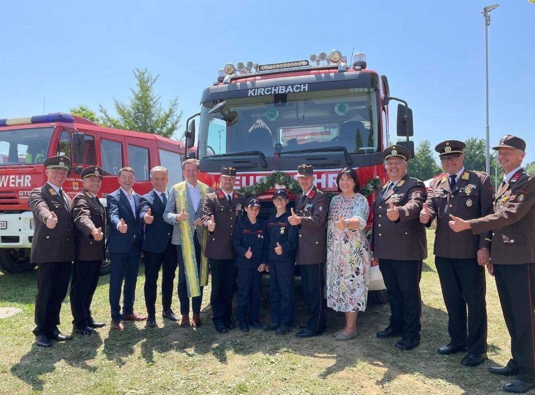 Eine Gruppe von Feuerwehrleuten und Beamten steht vor einem roten Feuerwehrauto namens Kirchbach. Dahinter steht ein weiteres Feuerwehrauto. Sie sind alle in Uniform gekleidet.