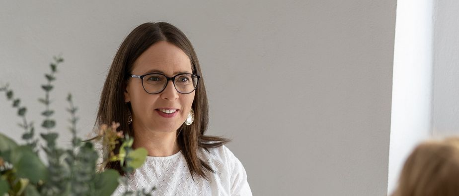 Eine Frau mit Brille und einer weißen Bluse sitzt an einem Schreibtisch, hält ein Buch und lächelt. Eine Vase mit weißen Blumen steht auf dem Schreibtisch. Eine andere Frau sitzt ihr gegenüber.
