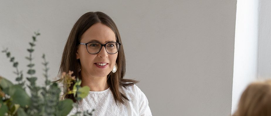 Eine Frau mit Brille und einer weißen Bluse sitzt an einem Tisch, lächelt und hält ein Buch. Ein Blumenstrauß mit weißen Blumen steht auf dem Tisch, und eine andere Frau sitzt ihr gegenüber.