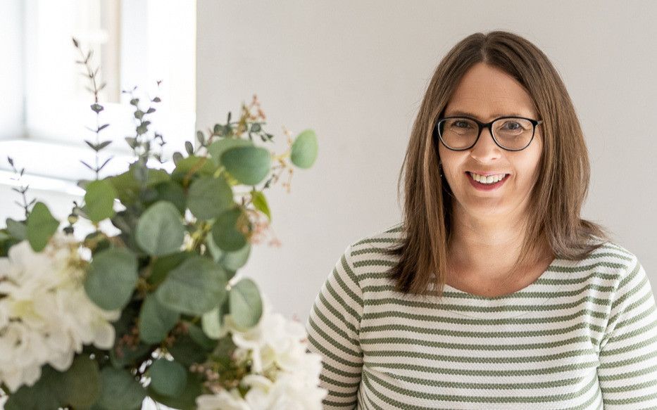 Eine Frau mit Brille lächelt neben einem Strauß grüner und weißer Blumen.