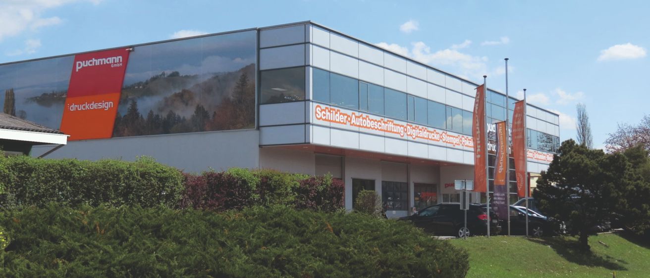 Ein modernes Gebäude mit einem großen Glasfenster, das eine Bergszene zeigt, befindet sich in einer sonnigen Umgebung. Vor dem Gebäude stehen Fahnen und ein geparktes Auto.