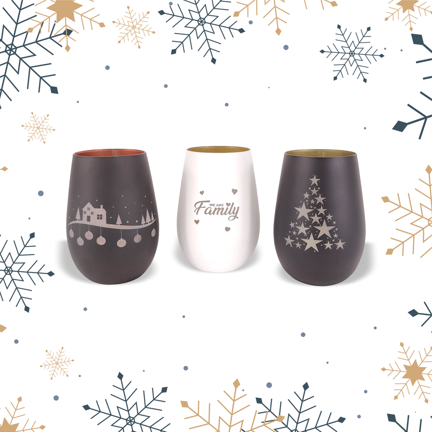 Drei Weingläser mit Weihnachtsdesigns auf weißem Hintergrund mit Schneeflocken und Sternen. Das linke Glas hat ein Haus- und Baumdesign, das mittlere Glas sagt 'We Are Family' und das rechte Glas hat ein Weihnachtsbaumdesign.