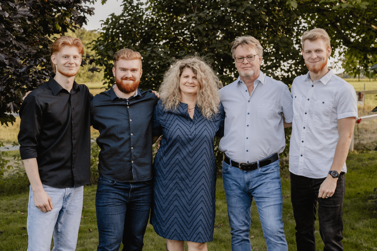 Fünf Personen posieren für ein Foto vor einer üppigen grünen Kulisse. Eine Frau mit lockigem Haar und einer Halskette steht in der Mitte, flankiert von zwei Männern auf jeder Seite.