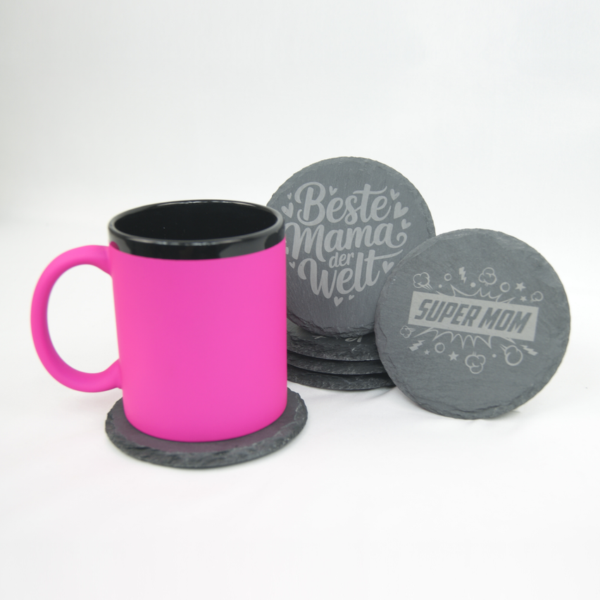 Eine rosa Kaffeetasse neben zwei Untersetzern, einer mit der Aufschrift 'Beste Mama der Welt' und der andere 'Super Mom'. Die Tasse und Untersetzer sind auf einer weißen Oberfläche.