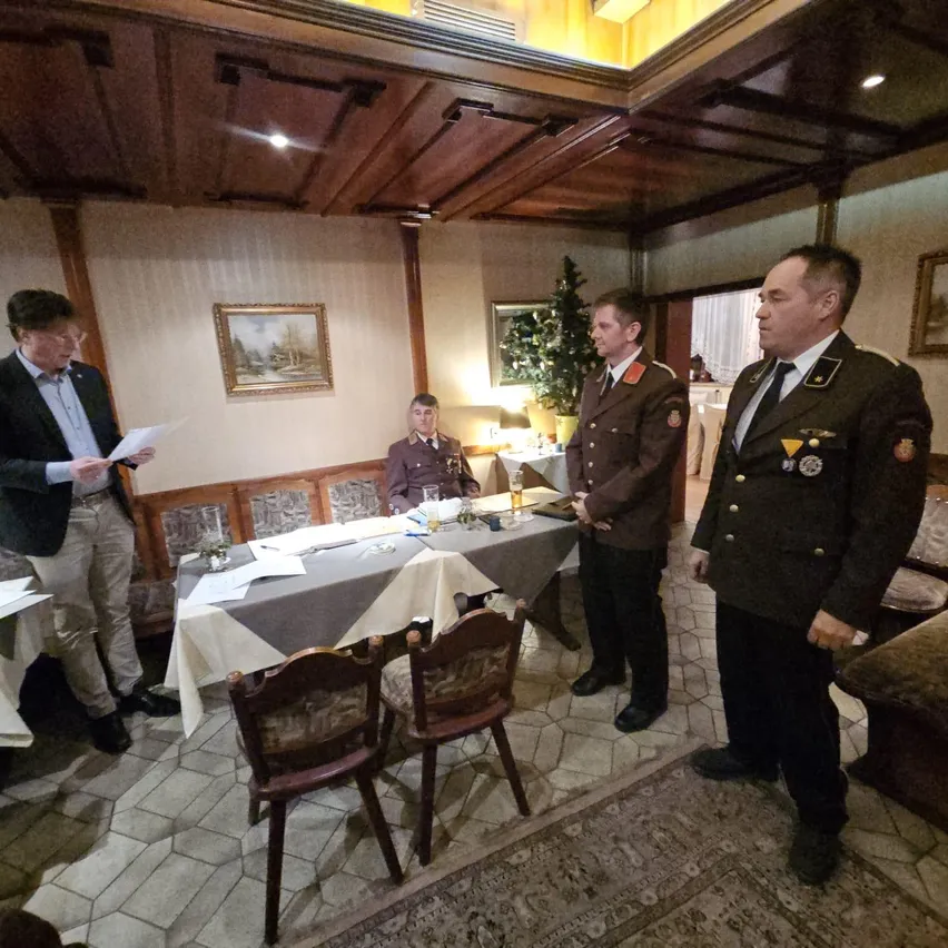 Drei Männer in Militäruniformen stehen um einen Tisch mit Papieren, während ein Mann in einem Anzug liest. Ein Weihnachtsbaum und Gemälde sind im Hintergrund.