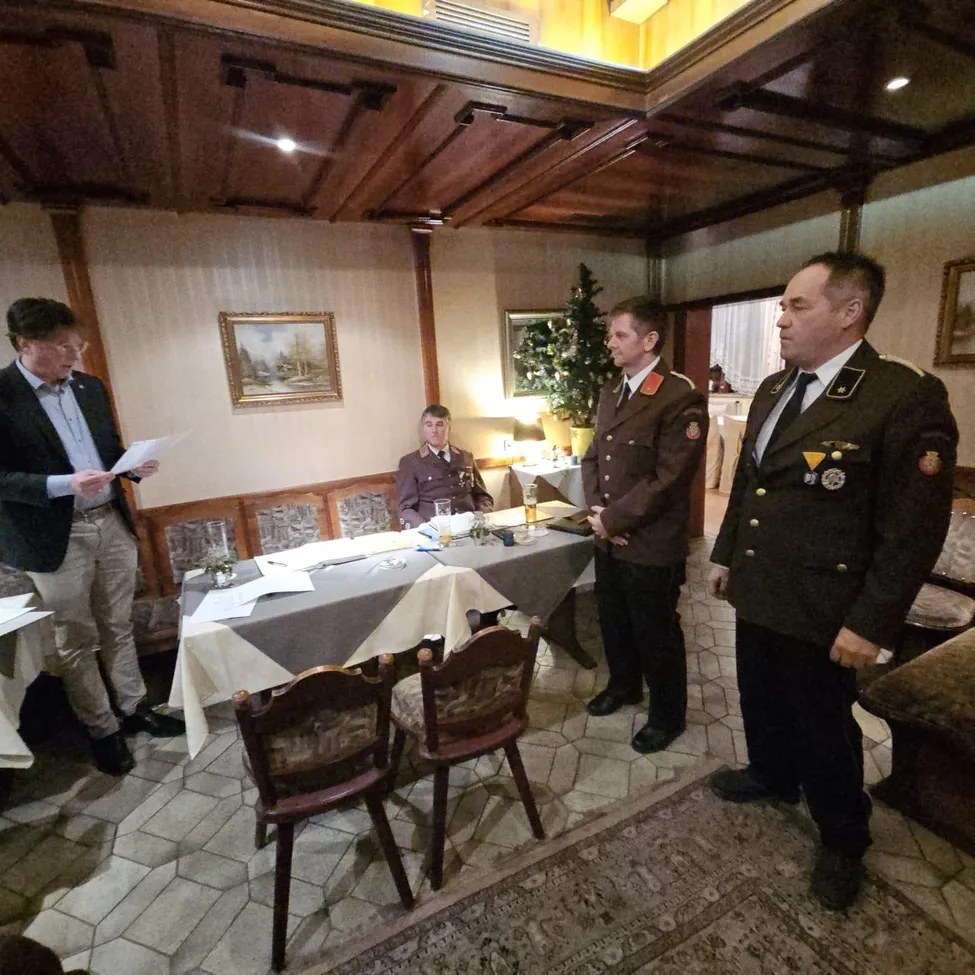 Drei Männer in Militäruniformen stehen um einen Tisch mit Papieren, während ein Mann in einem Anzug liest. Ein Weihnachtsbaum und Gemälde sind im Hintergrund.