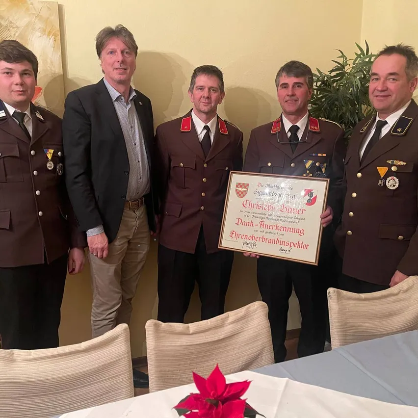Fünf Männer stehen in einem Raum, drei halten ein gerahmtes Zertifikat, einer lächelt und ein anderer hält ein gerahmtes Zertifikat. Der Mann in der Mitte trägt eine Medaille und eine Krawatte. Der Raum hat einen Tisch mit einer Blume und Stühlen.