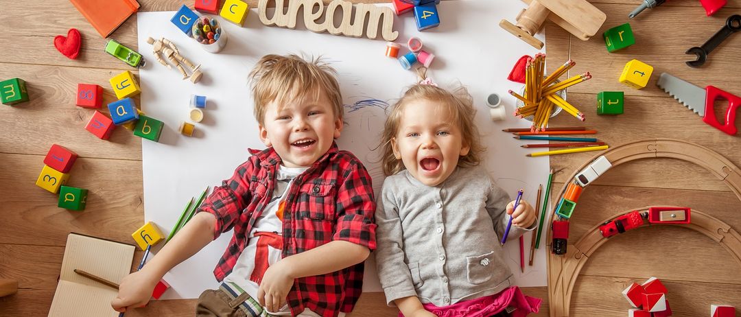 Zwei Kinder sitzen auf einem Holzboden, umgeben von Spielzeug, Büchern und Stiften. Der Junge lächelt und das Mädchen lacht. Das Wort 'Traum' ist in großen Holzbuchstaben auf dem Boden geschrieben.