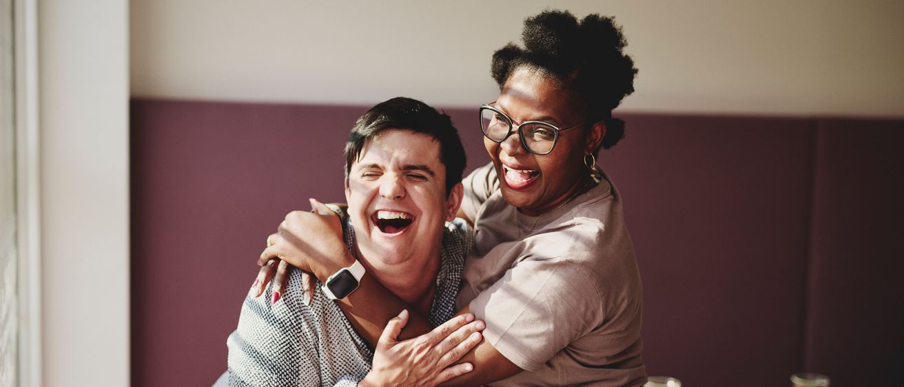 Zwei Menschen umarmen sich und lachen. Die Frau trägt eine Brille und Ohrringe, der Mann eine Uhr. Hinter ihnen steht ein Tisch mit einer Schüssel und einem Glas. Der Hintergrund besteht aus lila Wänden und einer Lampe.