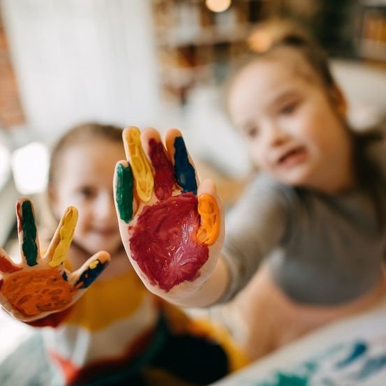 Zwei kleine Mädchen mit von Farbe bedeckten Händen schauen in die Kamera. Eines der Mädchen hat die Hände erhoben, das andere hat sie gesenkt.