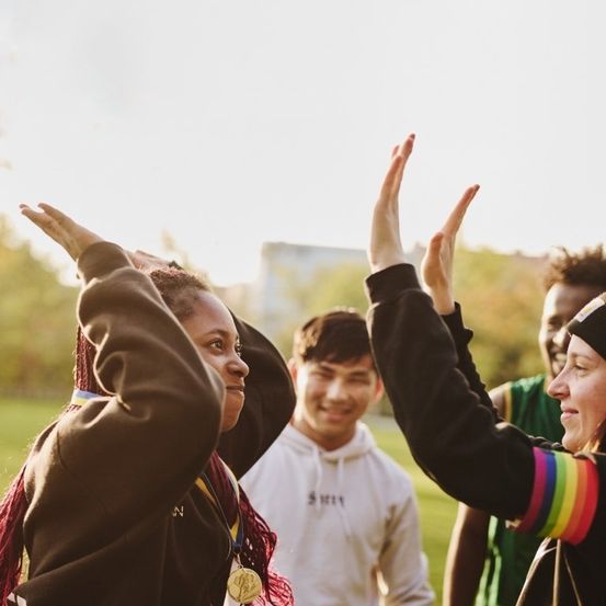 Eine Gruppe von Menschen ist draußen, zwei heben ihre Hände. Eine Frau hat Medaillen um den Hals. Hinter ihnen steht ein lächelnder Mann und in der Ferne ein Gebäude.